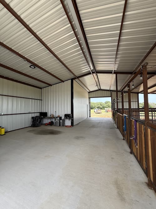 Barn interior breezeway with cedar stalls, lighting, and pasture beyond